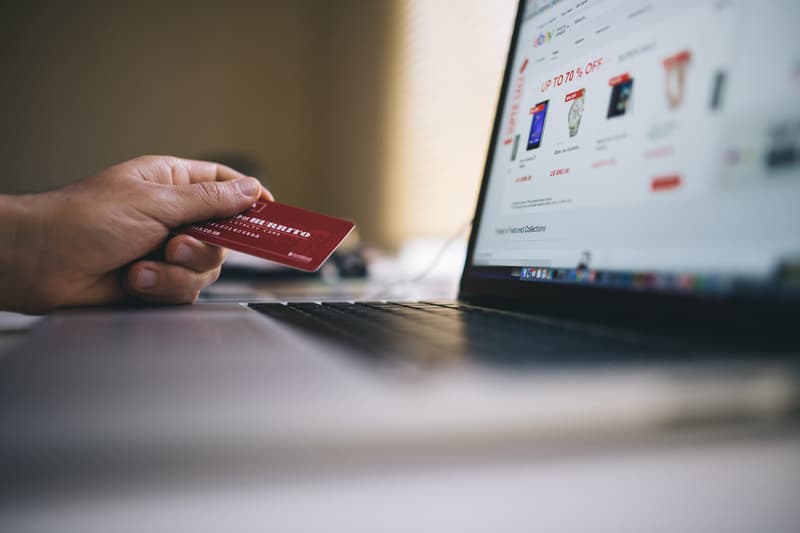 a hand holding a credit card in front of a laptop displaying products and "SALE 70% OFF" in bright red letters