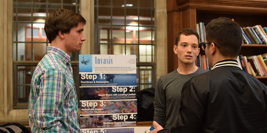 Two students explain their pitch idea to another student front of a posterboard in the Fulton Honors Library