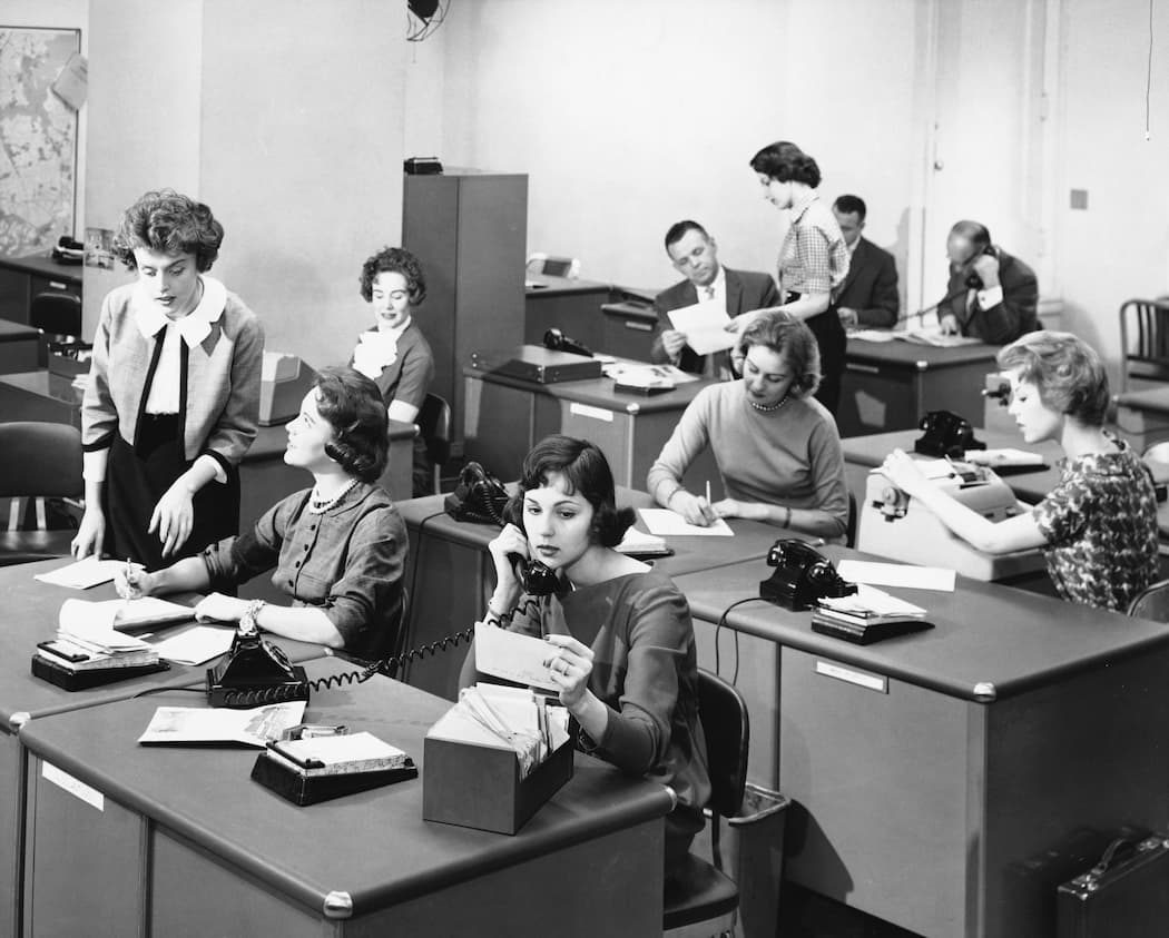 Men and women in a vintage office setting answering phones and filling out paperwork.