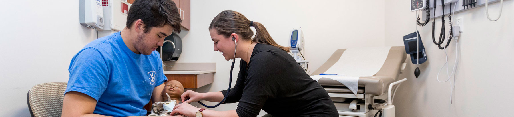 One student holds a medical mannikin of a baby, while a second student uses a stethoscope to listen to the heartbeat