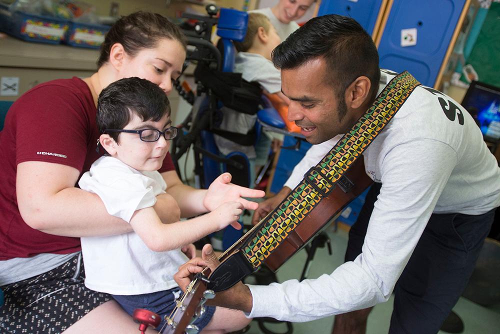 A young boy sitting on a teacher's lap watching a man play guitar