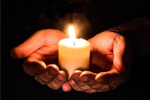 A pair of hands in darkness holding a lit votive candle