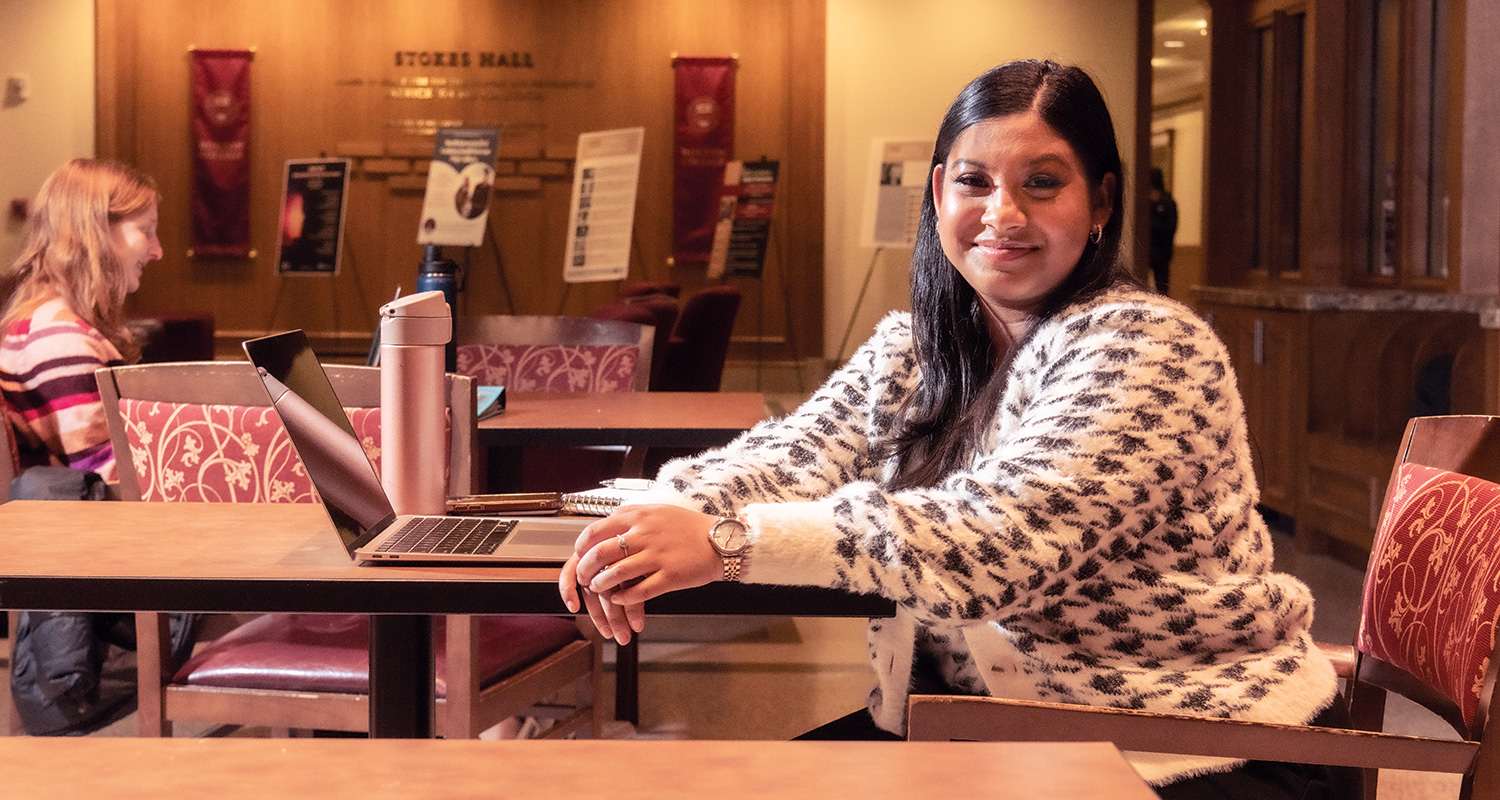 student in Bapst Library at a laptop, with other students at tables studying in the background