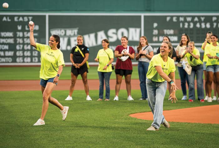 Two BC Lacrosse players throwing the first pitch with the team and coaching staff in the background