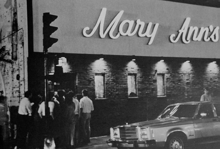 Black and white photograph of people gathered outside a bar in the evening