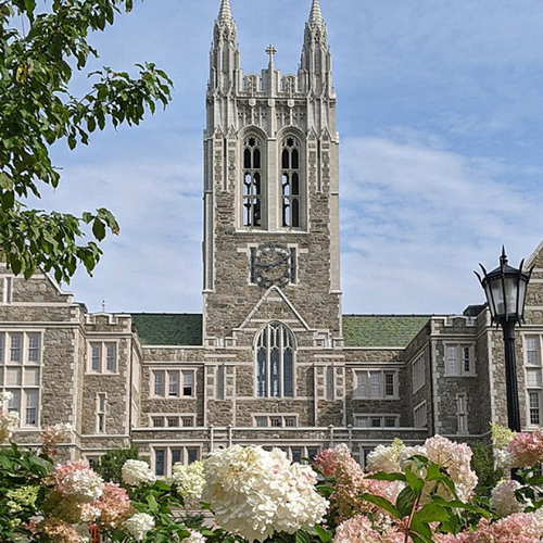 Gasson Hall, a stately gothic building, seen on a sunny day against a bright blue sky. Pink and white hydrangeas are visible in the foreground.