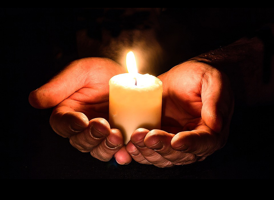 A pair of hands in darkeness holding a lit votive candle